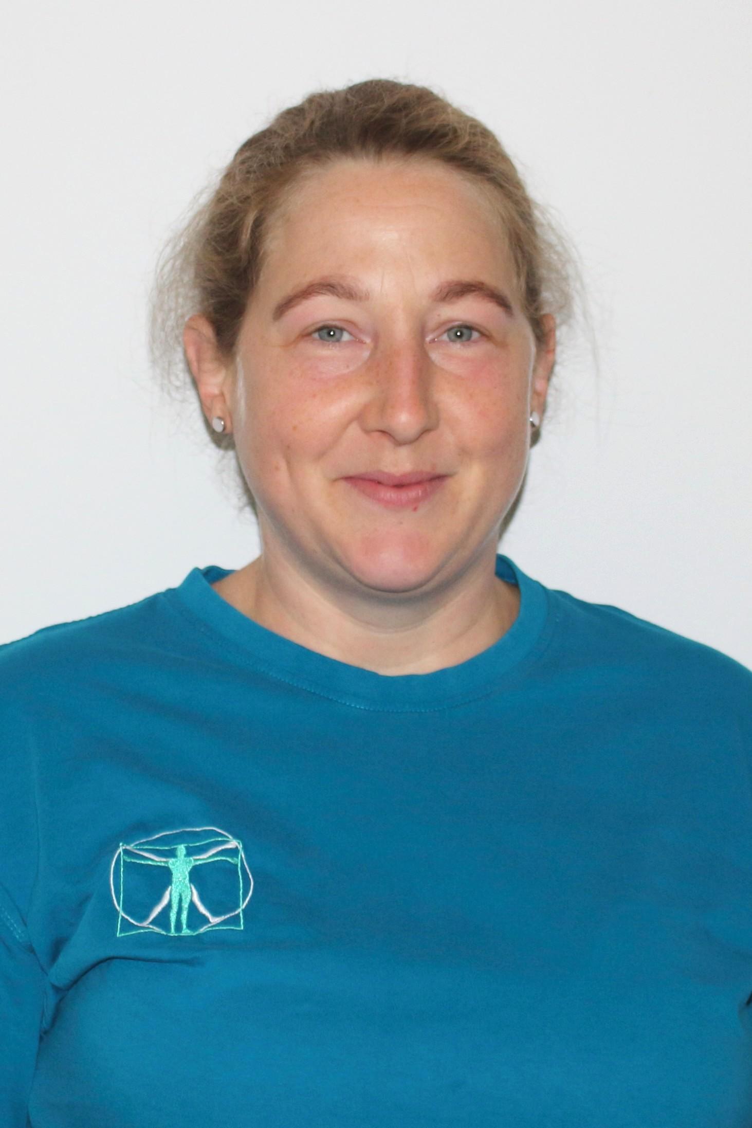 Eine Frau mit zurückgebundenen Haaren trägt ein türkisfarbenes Oberteil mit einem Vitruvianischen Mensch Motiv und schaut direkt in die Kamera, während sie leicht lächelt.